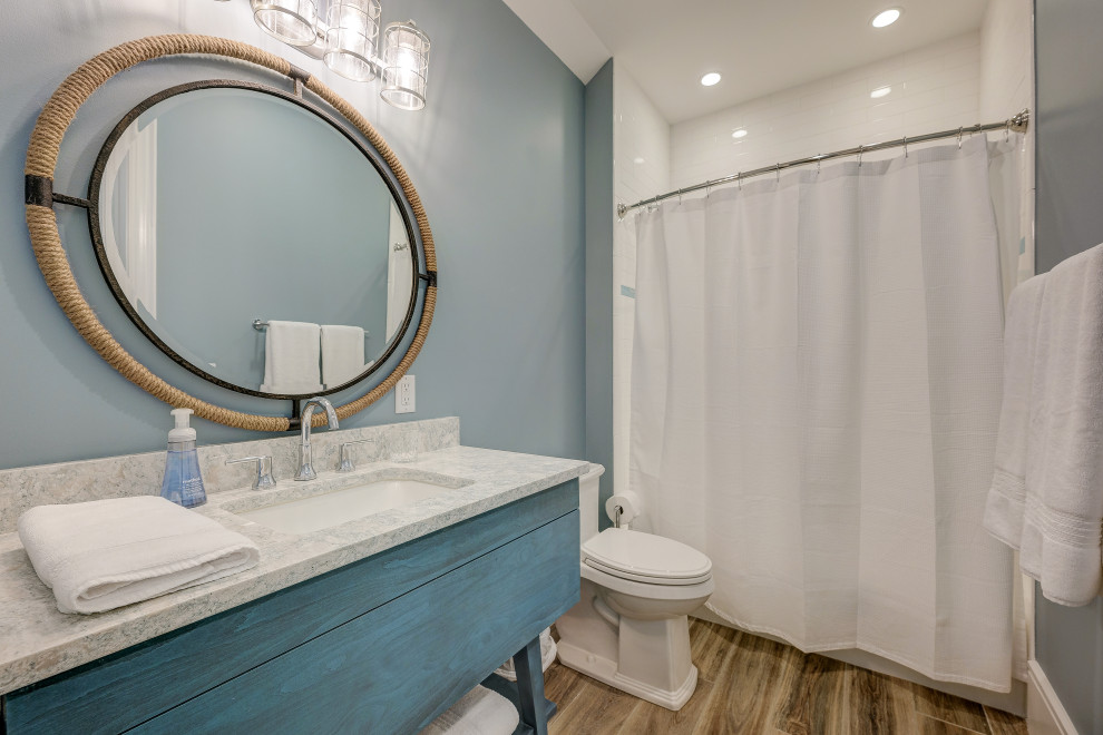 This is an example of a mid-sized beach style kids bathroom in Other with blue cabinets, a shower/bathtub combo, white tile, porcelain tile, blue walls, an undermount sink, marble benchtops, brown floor, a shower curtain, white benchtops, a single vanity, a freestanding vanity, furniture-like cabinets, an alcove tub, a one-piece toilet and medium hardwood floors.