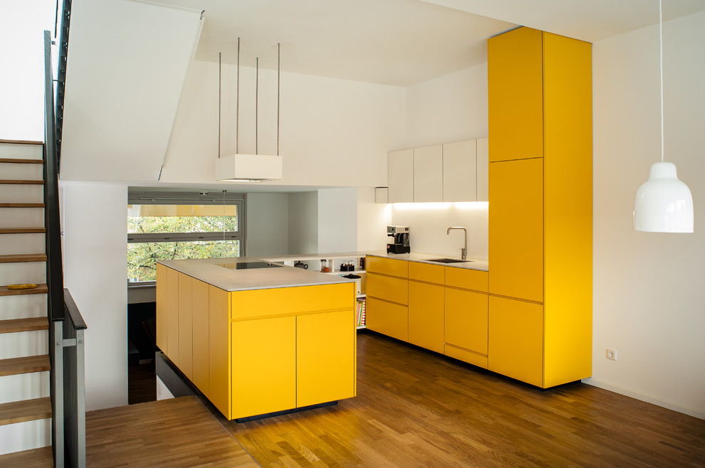 This is an example of a mid-sized modern u-shaped open plan kitchen in Berlin with an integrated sink, flat-panel cabinets, yellow cabinets, white splashback, panelled appliances, medium hardwood floors, a peninsula, brown floor and grey benchtop.