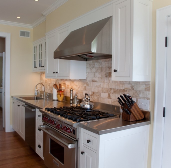 This is an example of a traditional l-shaped eat-in kitchen in Burlington with an undermount sink, shaker cabinets, white cabinets, stainless steel benchtops, beige splashback, stone tile splashback, stainless steel appliances and medium hardwood floors.