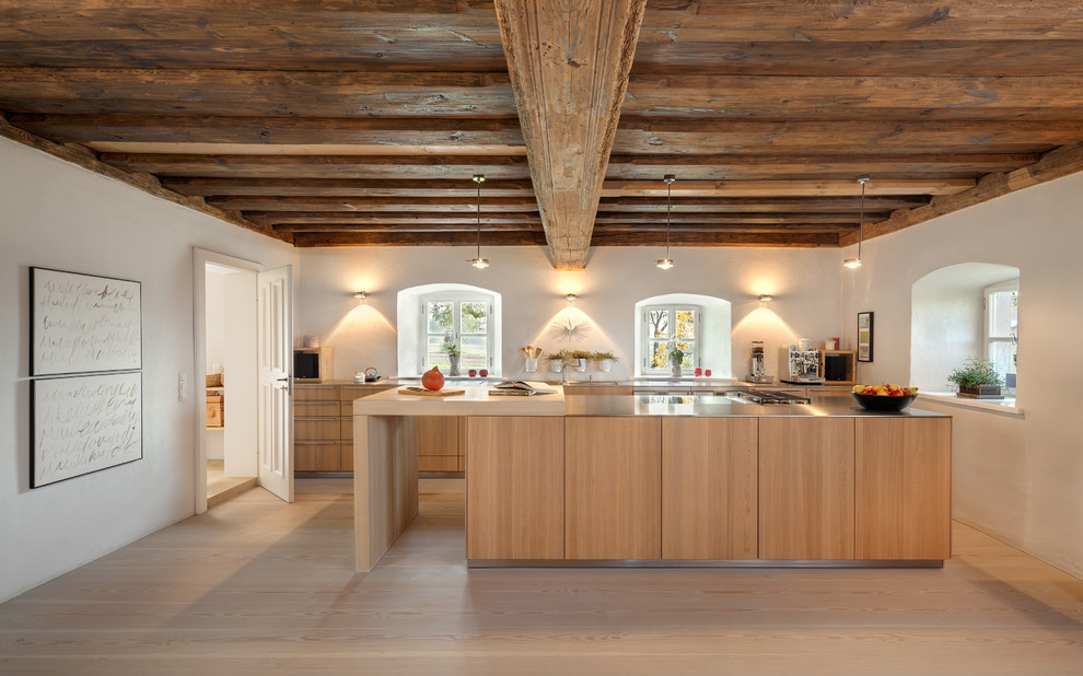 This is an example of a large contemporary galley open plan kitchen in Nuremberg with flat-panel cabinets, light wood cabinets, stainless steel benchtops, white splashback, light hardwood floors and with island.