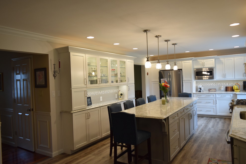 Example of a huge transitional medium tone wood floor and brown floor kitchen design with an undermount sink, shaker cabinets, white cabinets, quartz countertops, gray backsplash, subway tile backsplash, stainless steel appliances and an island