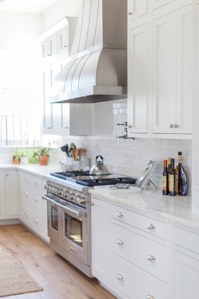 Inspiration for a large transitional l-shaped light wood floor eat-in kitchen remodel in Phoenix with a farmhouse sink, white cabinets, white backsplash, stainless steel appliances, an island, shaker cabinets, stainless steel countertops and porcelain backsplash