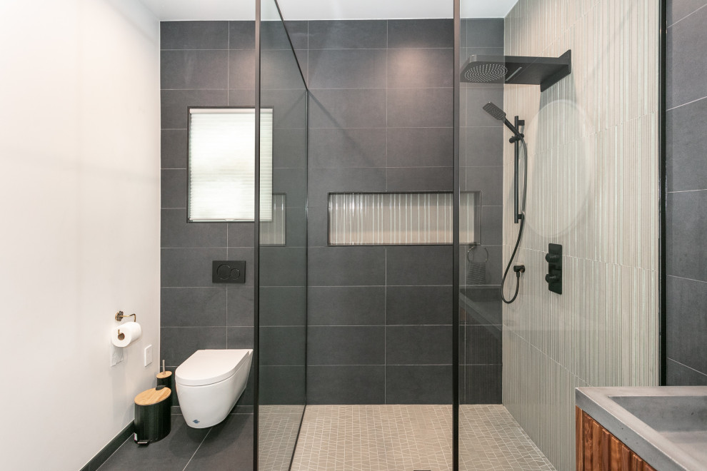 Bild på ett mellanstort badrum, med skåp i mörkt trä, en hörndusch, en vägghängd toalettstol, grå kakel, porslinskakel, vita väggar, klinkergolv i porslin, ett avlångt handfat, grått golv och med dusch som är öppen