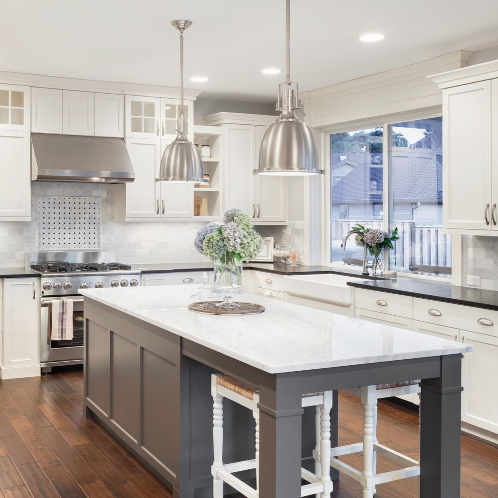 This is an example of a transitional l-shaped kitchen in Toronto with a farmhouse sink, shaker cabinets, white cabinets, multi-coloured splashback, stainless steel appliances, dark hardwood floors, with island, brown floor and black benchtop.
