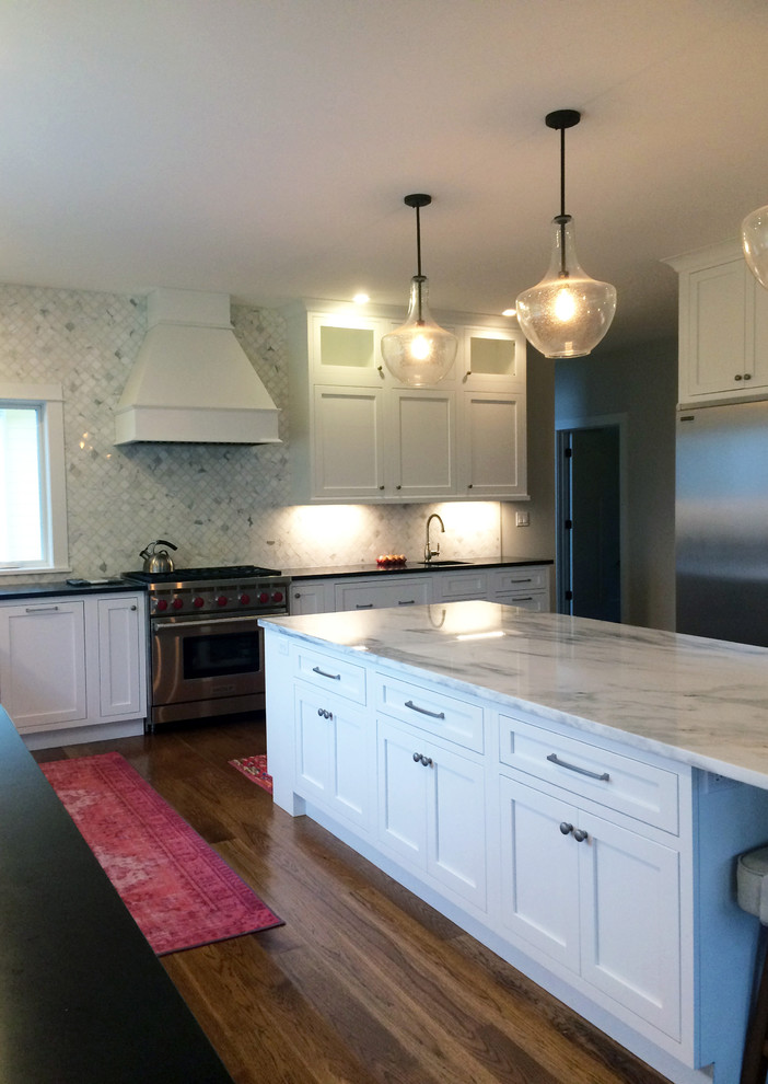 Example of a large transitional l-shaped dark wood floor and brown floor kitchen pantry design with a farmhouse sink, recessed-panel cabinets, white cabinets, quartz countertops, beige backsplash, ceramic backsplash, stainless steel appliances, an island and black countertops