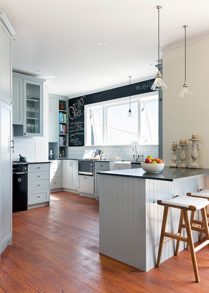 This is an example of a transitional u-shaped kitchen in Sydney with a farmhouse sink, shaker cabinets, grey cabinets, white splashback, subway tile splashback, black appliances, dark hardwood floors, a peninsula, brown floor and black benchtop.