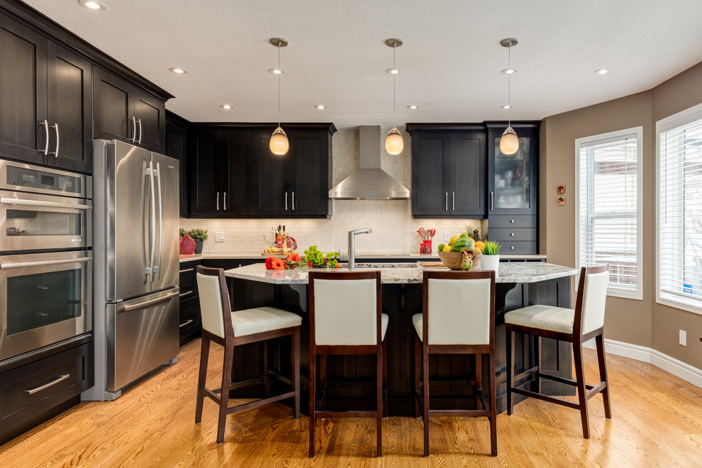 Transitional l-shaped kitchen in Calgary with an undermount sink, shaker cabinets, black cabinets, white splashback, stainless steel appliances, medium hardwood floors and with island.