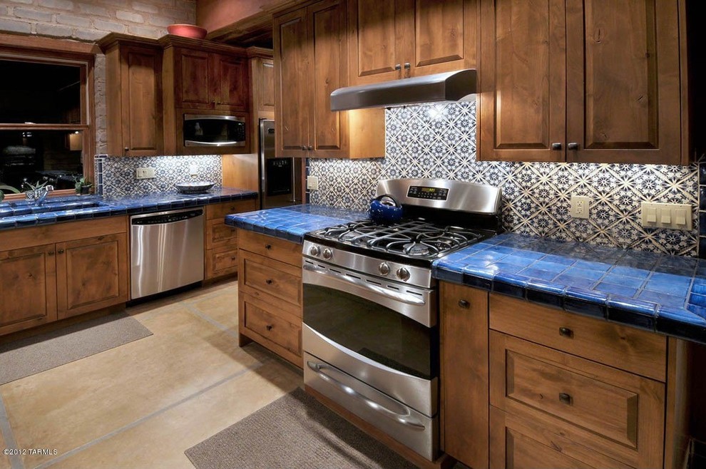 Photo of a mid-sized l-shaped eat-in kitchen in Phoenix with a double-bowl sink, raised-panel cabinets, medium wood cabinets, tile benchtops, blue splashback, ceramic splashback, stainless steel appliances, ceramic floors and with island.