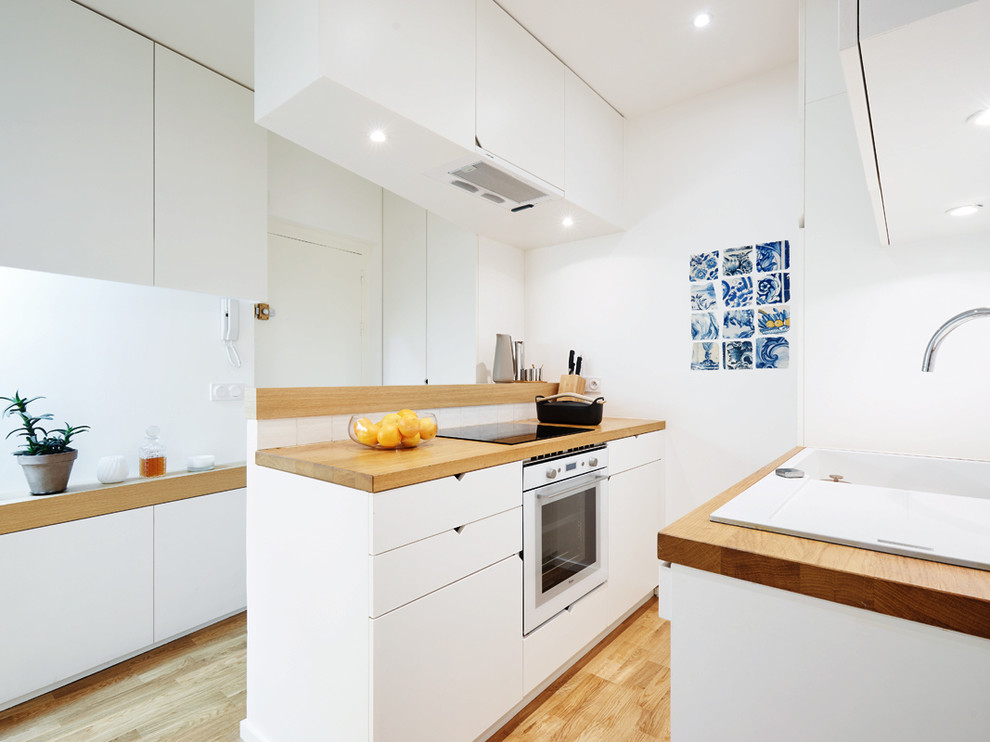 Photo of a small contemporary galley open plan kitchen in Paris with flat-panel cabinets, white cabinets, wood benchtops, white appliances, light hardwood floors, an undermount sink and no island.