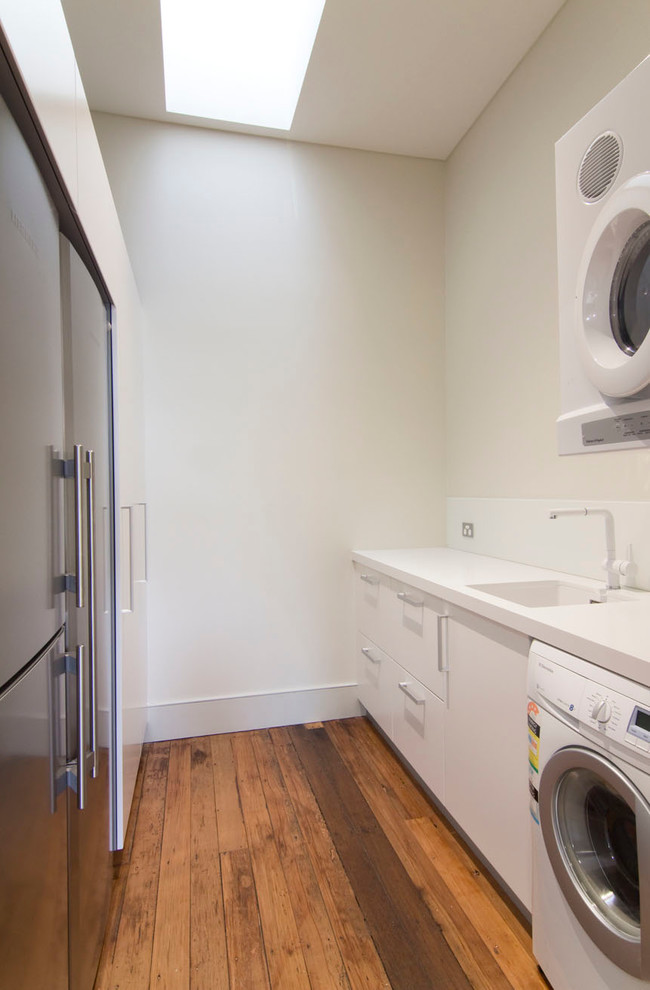 Photo of a small modern galley utility room in Sydney with an undermount sink, flat-panel cabinets, quartz benchtops, white splashback, glass sheet splashback, white cabinets, white walls, medium hardwood floors and a stacked washer and dryer.