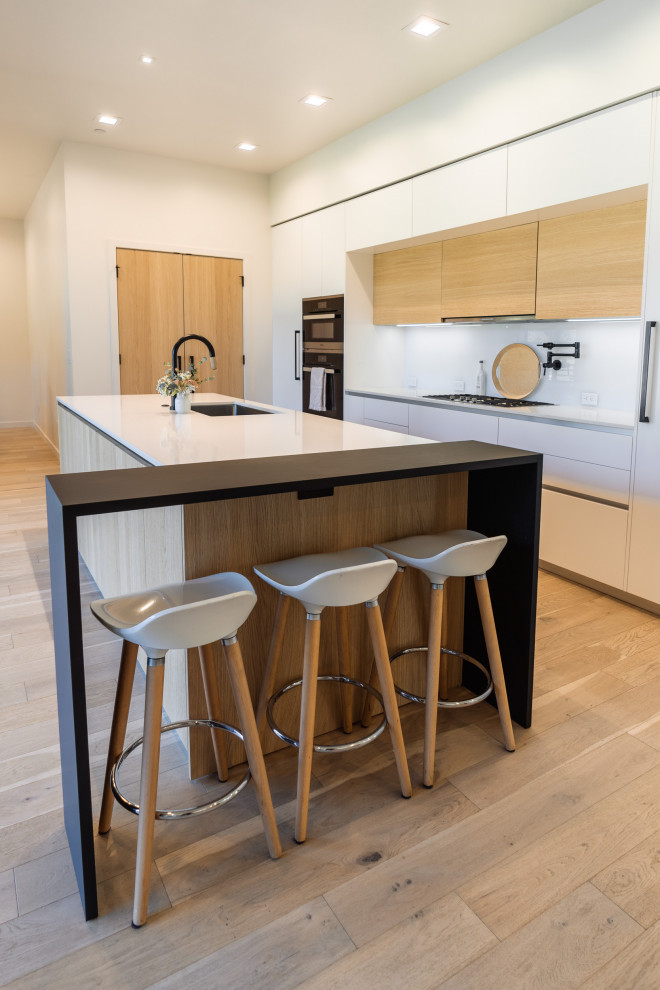 Expansive modern single-wall eat-in kitchen in Portland with an undermount sink, flat-panel cabinets, light wood cabinets, quartz benchtops, white splashback, glass sheet splashback, panelled appliances, light hardwood floors, with island, brown floor and white benchtop.