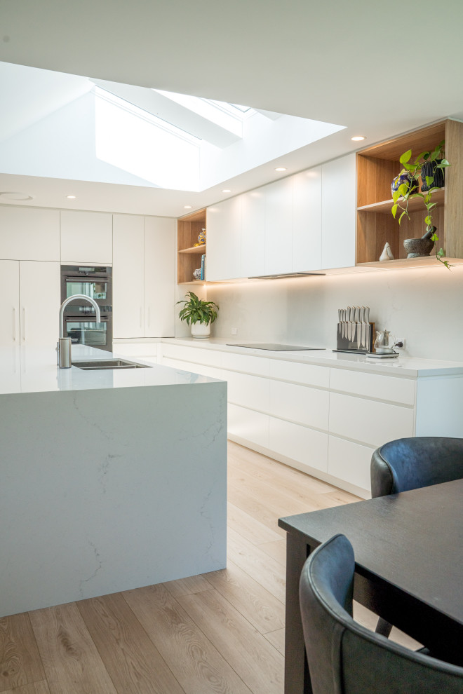 Photo of a large modern l-shaped open plan kitchen in Sydney with a double-bowl sink, flat-panel cabinets, white cabinets, engineered stone countertops, white splashback, engineered quartz splashback, black appliances, laminate floors, an island, brown floors and white worktops.