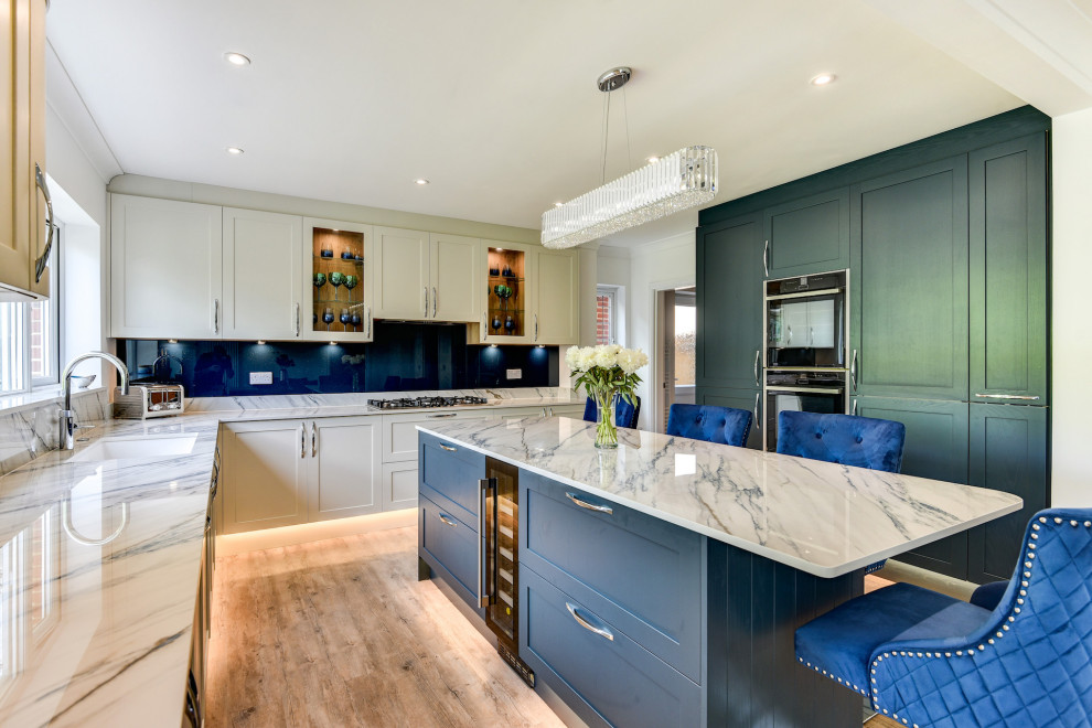 Medium sized classic l-shaped kitchen/diner in Sussex with a submerged sink, shaker cabinets, blue cabinets, composite countertops, blue splashback, glass sheet splashback, black appliances, medium hardwood flooring, an island, brown floors, white worktops and feature lighting.