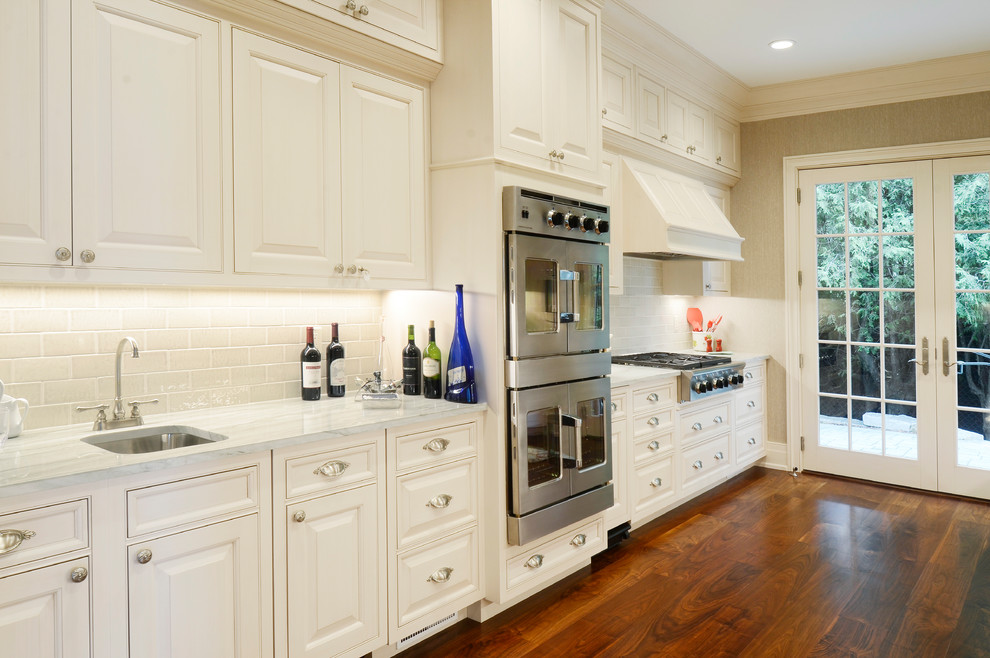 This is an example of an expansive traditional u-shaped separate kitchen in Other with an undermount sink, raised-panel cabinets, white cabinets, marble benchtops, porcelain splashback, stainless steel appliances, medium hardwood floors, multiple islands, brown floor, white benchtop and white splashback.