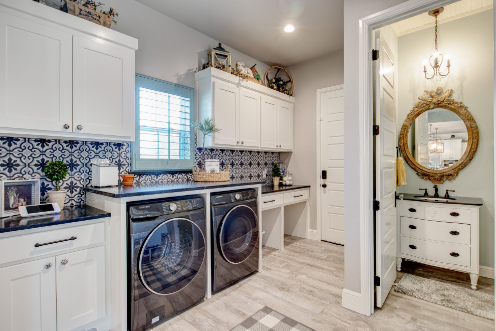 This is an example of a mid-sized country single-wall utility room in Dallas with shaker cabinets, white cabinets, granite benchtops, black splashback, ceramic splashback, grey walls, ceramic floors, a side-by-side washer and dryer, grey floor and black benchtop.