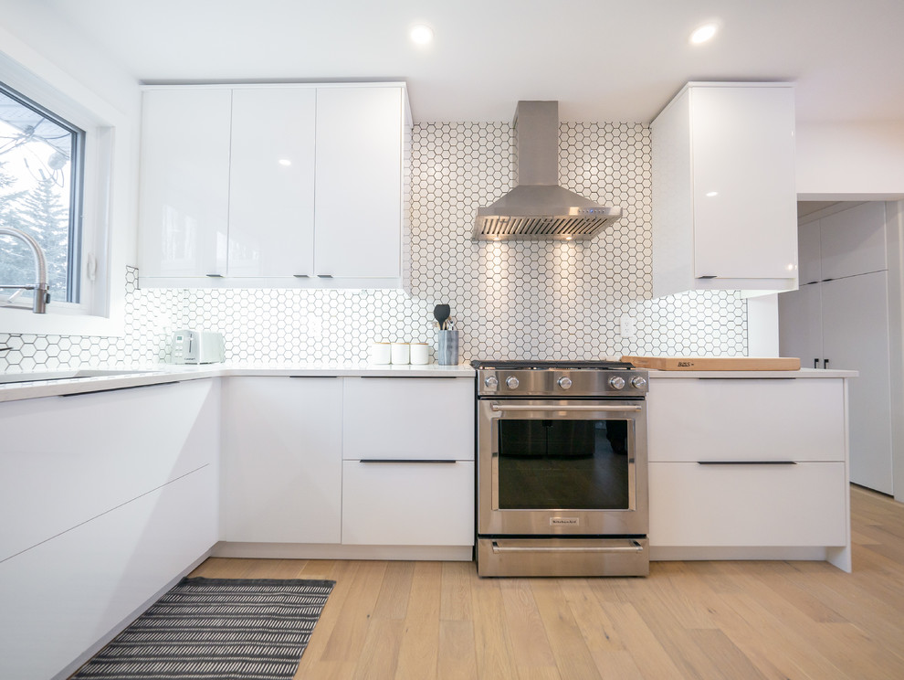 This is an example of a mid-sized midcentury l-shaped kitchen in Edmonton with white cabinets, white splashback, stainless steel appliances, white benchtop, an undermount sink, flat-panel cabinets, solid surface benchtops, light hardwood floors, a peninsula, brown floor and porcelain splashback.