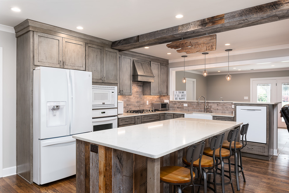 Design ideas for a large country l-shaped open plan kitchen in Other with a farmhouse sink, shaker cabinets, medium wood cabinets, quartz benchtops, grey splashback, brick splashback, white appliances, dark hardwood floors, with island, brown floor and white benchtop.