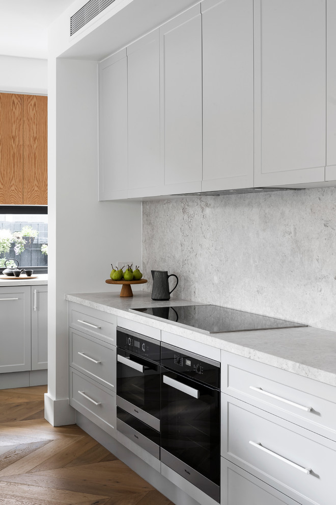 This is an example of a contemporary galley open plan kitchen in Melbourne with a farmhouse sink, shaker cabinets, grey cabinets, limestone benchtops, grey splashback, limestone splashback, black appliances, medium hardwood floors, with island, brown floor and grey benchtop.