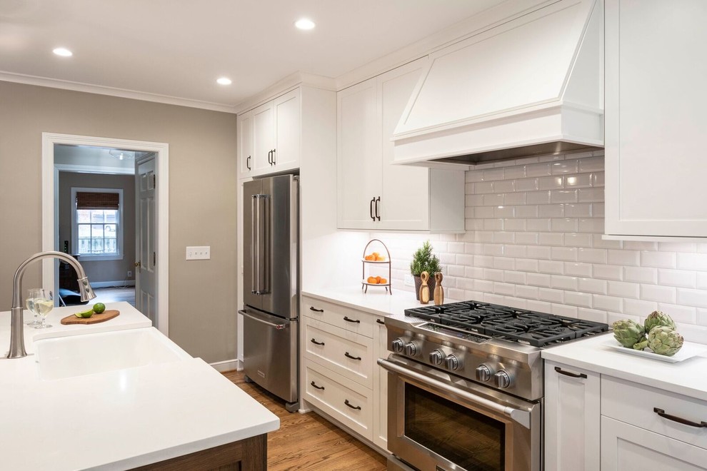 This is an example of a mid-sized transitional l-shaped eat-in kitchen in Indianapolis with a farmhouse sink, shaker cabinets, white cabinets, quartz benchtops, white splashback, ceramic splashback, stainless steel appliances, medium hardwood floors, with island, brown floor and white benchtop.