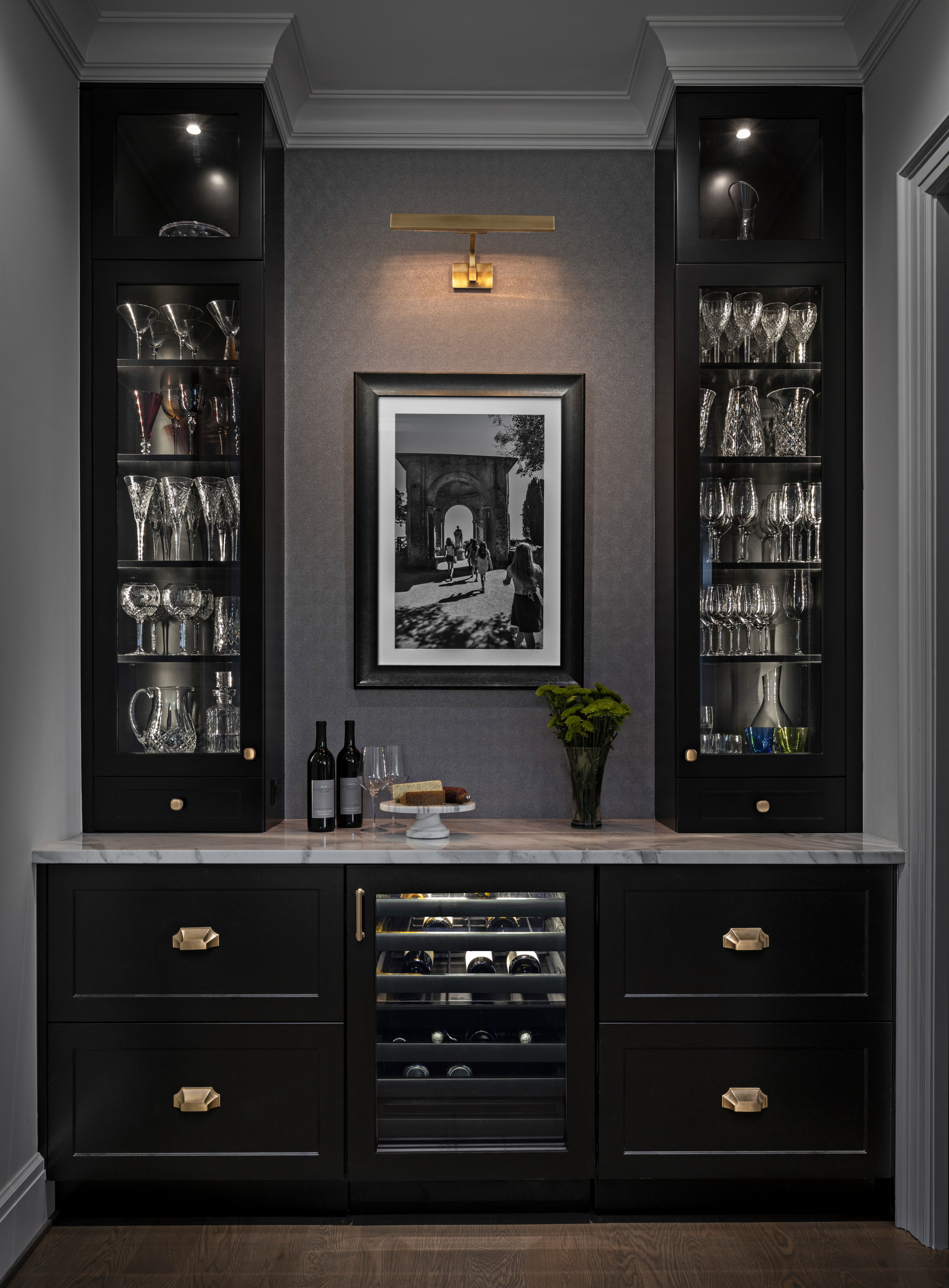 What makes a smaller space shine? In this Butler’s Pantry, our gorgeous dark shaker cabinets frame the view. Wallpaper with texture and a picture light play off the dramatic hardware to give some glam