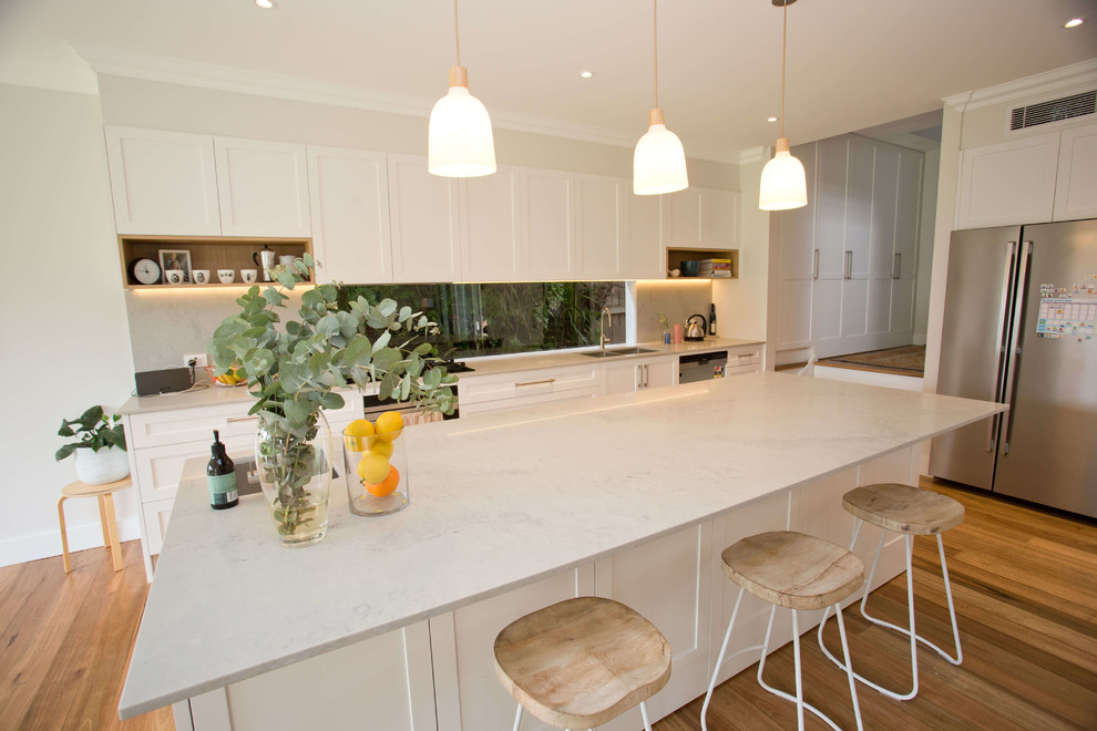 This is an example of a transitional l-shaped eat-in kitchen in Sydney with a double-bowl sink, shaker cabinets, white cabinets, window splashback, stainless steel appliances, dark hardwood floors, with island and beige benchtop.