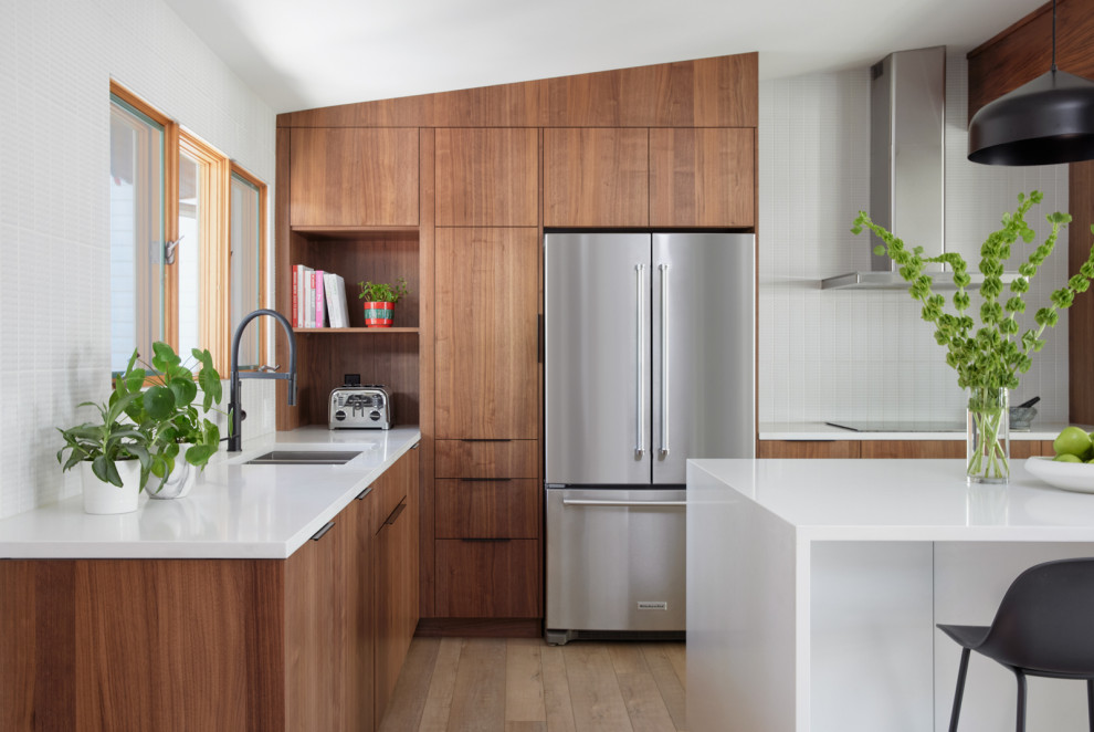 This is an example of a mid-sized midcentury l-shaped eat-in kitchen in Calgary with a double-bowl sink, flat-panel cabinets, medium wood cabinets, quartzite benchtops, white splashback, ceramic splashback, stainless steel appliances, vinyl floors, with island, brown floor, white benchtop and vaulted.