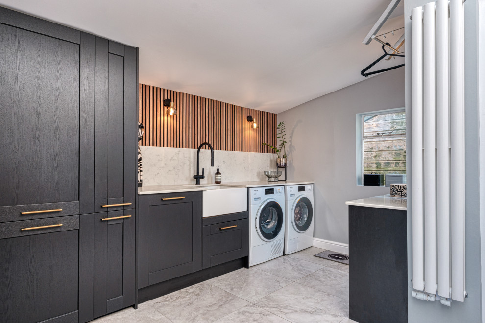 Photo of a large contemporary l-shaped laundry room in London with a farmhouse sink, shaker cabinets, black cabinets, white splashback, engineered quartz splashback, grey walls, porcelain floors, a side-by-side washer and dryer, grey floor, white benchtop, recessed, panelled walls and quartzite benchtops.