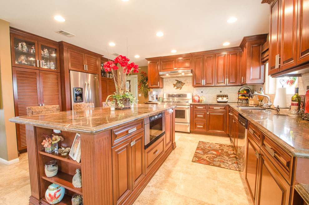 Large tropical galley separate kitchen in Orange County with a drop-in sink, raised-panel cabinets, dark wood cabinets, granite benchtops, white splashback, ceramic splashback, stainless steel appliances, travertine floors and with island.