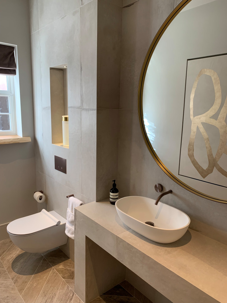 Inspiration for a mid-sized contemporary powder room in Cheshire with flat-panel cabinets, a wall-mount toilet, gray tile, ceramic tile, white walls, ceramic floors, a trough sink, tile benchtops, grey floor and white benchtops.