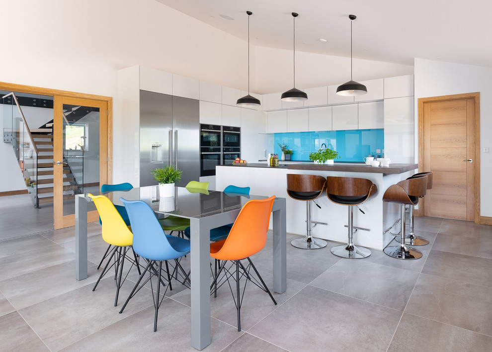 Photo of a large contemporary l-shaped open plan kitchen in Gloucestershire with an integrated sink, flat-panel cabinets, white cabinets, solid surface benchtops, blue splashback, glass sheet splashback, black appliances, porcelain floors, with island, grey floor and brown benchtop.