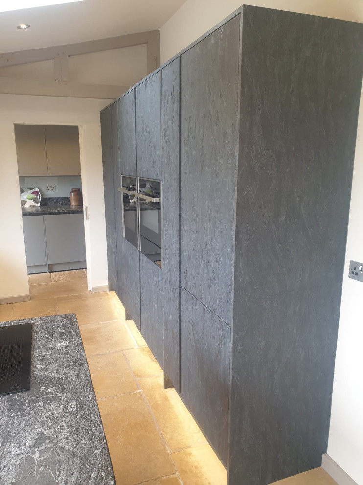 This is an example of a medium sized contemporary grey and black l-shaped kitchen/diner in West Midlands with flat-panel cabinets, granite worktops, black splashback, engineered quartz splashback, stainless steel appliances, black worktops, feature lighting, a built-in sink, black cabinets, travertine flooring, a breakfast bar, beige floors and a vaulted ceiling.