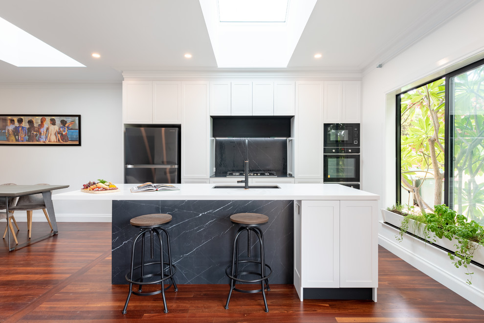 This is an example of a mid-sized transitional galley open plan kitchen in Perth with an undermount sink, shaker cabinets, quartz benchtops, grey splashback, stone slab splashback, black appliances, medium hardwood floors, with island, white benchtop and brown floor.