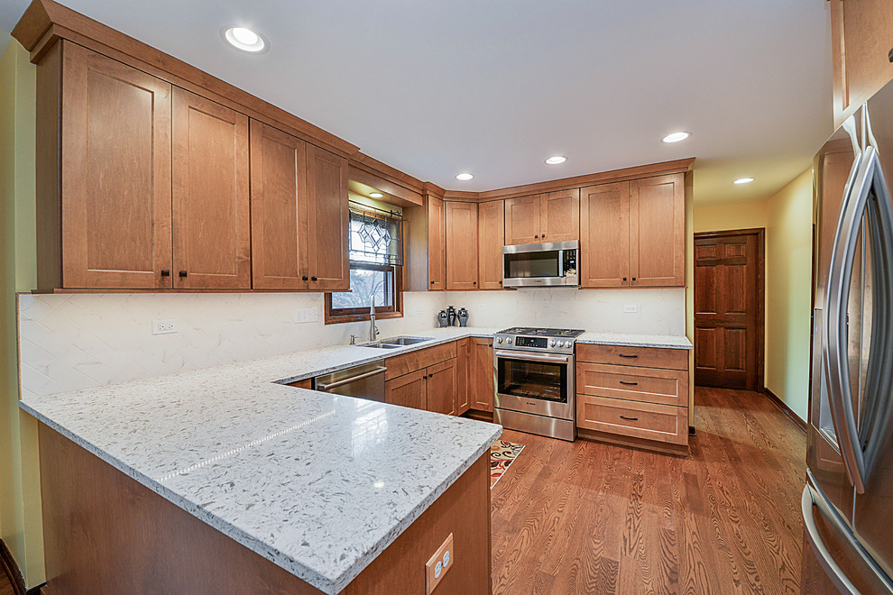 This is an example of a mid-sized transitional u-shaped eat-in kitchen in Chicago with an undermount sink, shaker cabinets, medium wood cabinets, quartz benchtops, white splashback, terra-cotta splashback, stainless steel appliances, medium hardwood floors and a peninsula.