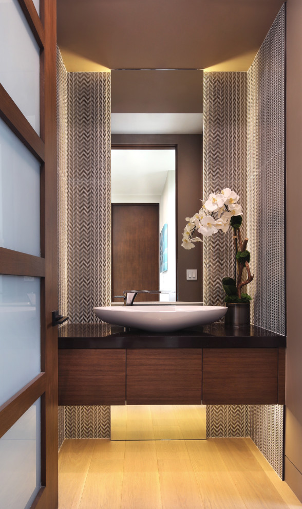 Inspiration for a small contemporary cloakroom in Los Angeles with flat-panel cabinets, brown cabinets, a one-piece toilet, grey tiles, mosaic tiles, brown walls, light hardwood flooring, a vessel sink, engineered stone worktops, beige floors, black worktops, feature lighting and a floating vanity unit.