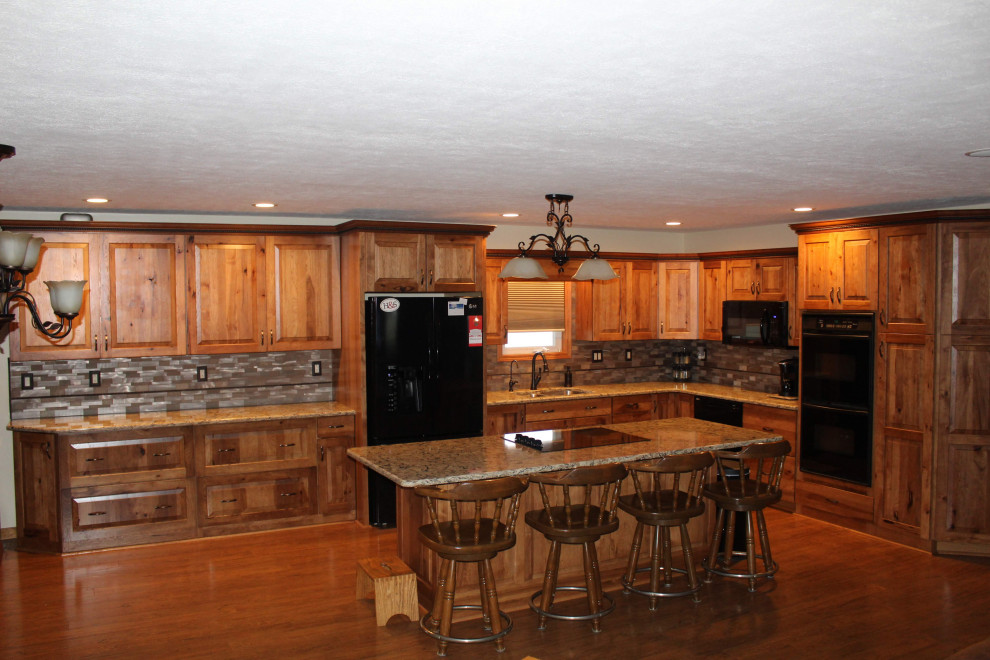 Example of a large mountain style l-shaped medium tone wood floor and brown floor open concept kitchen design with an undermount sink, raised-panel cabinets, brown cabinets, quartzite countertops, metallic backsplash, black appliances, an island and brown countertops