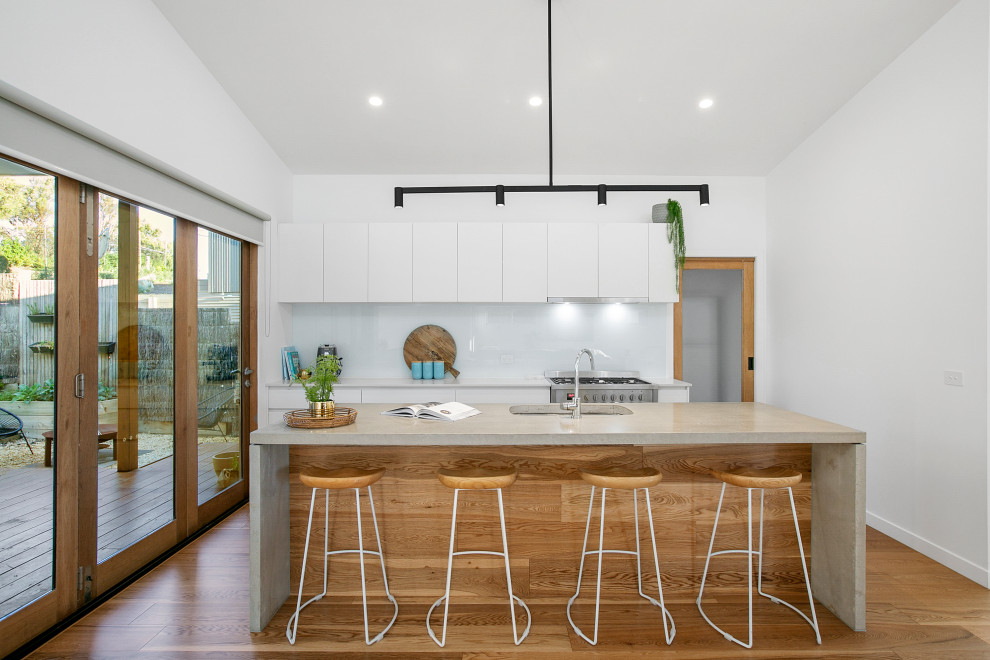 Mid-sized contemporary galley open plan kitchen in Melbourne with an undermount sink, flat-panel cabinets, white cabinets, concrete benchtops, white splashback, glass sheet splashback, stainless steel appliances, medium hardwood floors, with island, beige floor, grey benchtop and vaulted.