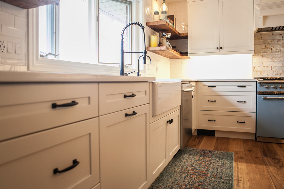 Large country u-shaped kitchen in Toronto with a farmhouse sink, shaker cabinets, white cabinets, solid surface benchtops, white splashback, porcelain splashback, coloured appliances, light hardwood floors, with island, brown floor and white benchtop.