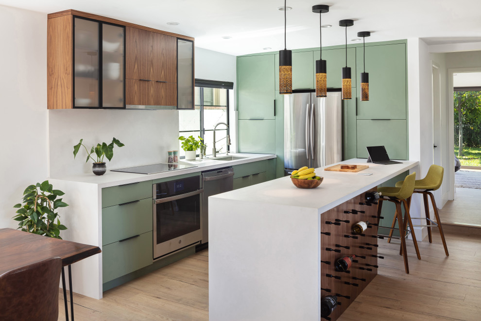 This is an example of a small contemporary galley open plan kitchen in Los Angeles with a submerged sink, flat-panel cabinets, green cabinets, engineered stone countertops, white splashback, engineered quartz splashback, stainless steel appliances, light hardwood flooring, a breakfast bar, brown floors and white worktops.