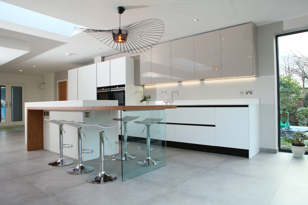 This is an example of a large contemporary single-wall open plan kitchen in West Midlands with a drop-in sink, flat-panel cabinets, white cabinets, solid surface benchtops, black appliances, cement tiles, with island and grey floor.