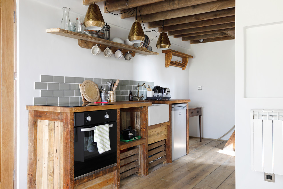 Photo of a small country single-wall open plan kitchen in Sussex with a farmhouse sink, open cabinets, distressed cabinets, wood benchtops, grey splashback, subway tile splashback, black appliances, medium hardwood floors and no island.
