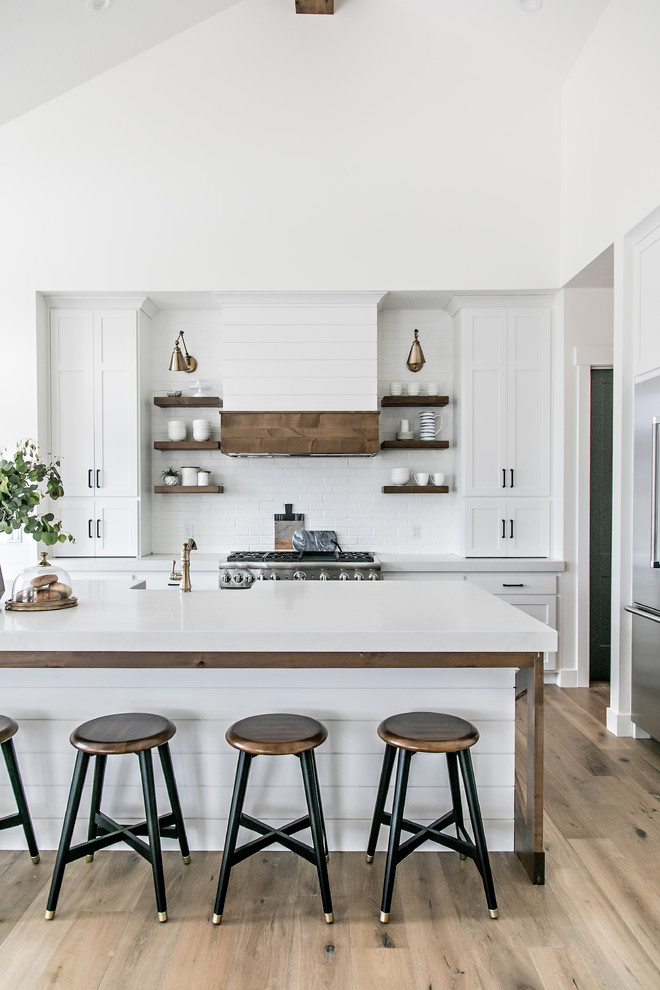 Inspiration for a mid-sized country l-shaped eat-in kitchen in Salt Lake City with a farmhouse sink, shaker cabinets, white cabinets, quartz benchtops, white splashback, subway tile splashback, stainless steel appliances, medium hardwood floors, with island, brown floor and white benchtop.