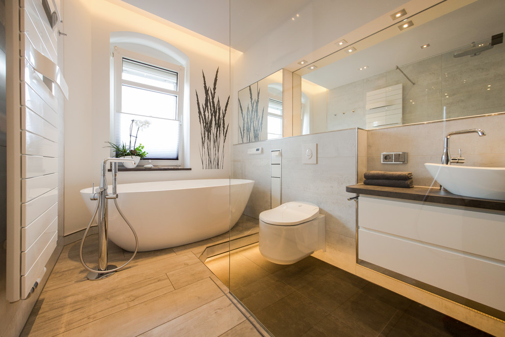 Design ideas for a mid-sized contemporary master bathroom in Dresden with flat-panel cabinets, white cabinets, a freestanding tub, a curbless shower, a two-piece toilet, beige tile, white walls, a vessel sink, beige floor and black benchtops.