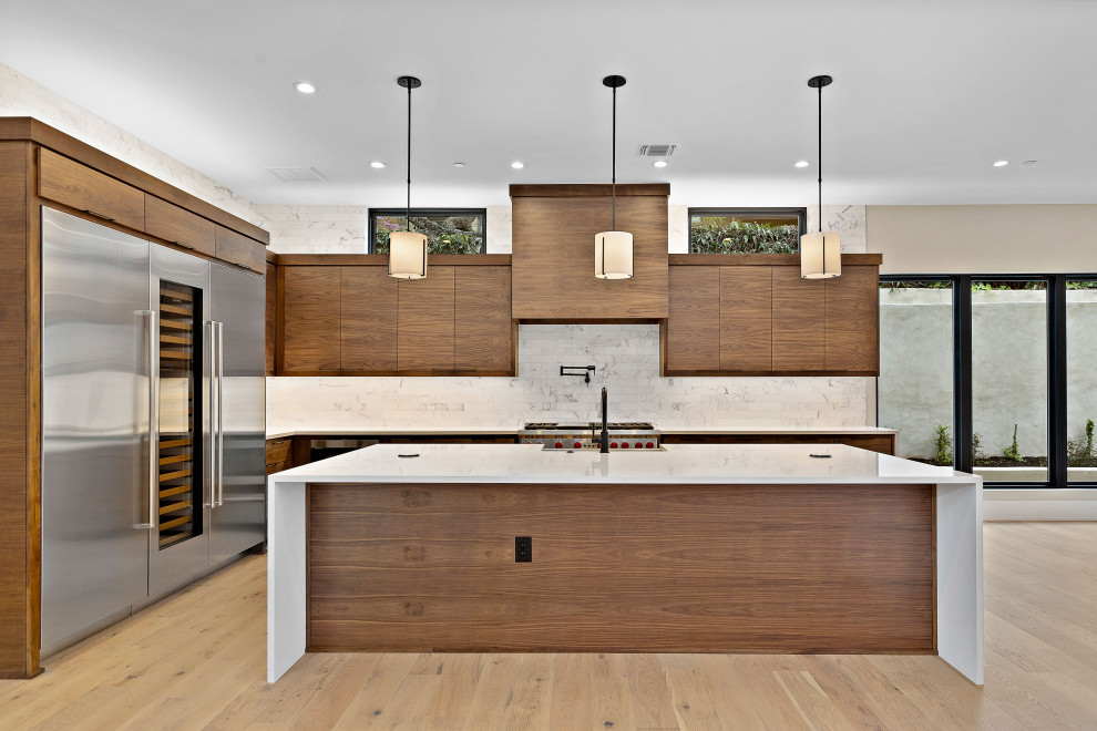 Photo of a large contemporary l-shaped eat-in kitchen in Austin with a farmhouse sink, flat-panel cabinets, medium wood cabinets, quartz benchtops, white splashback, marble splashback, stainless steel appliances, light hardwood floors, with island and white benchtop.