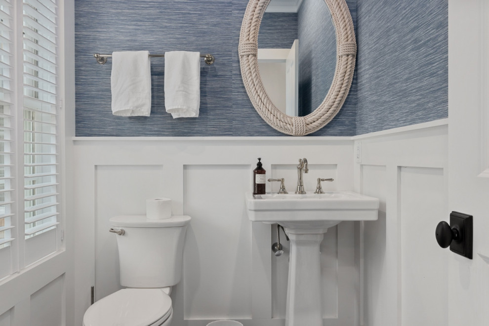 Beach style powder room in Boston with a two-piece toilet, blue walls, a pedestal sink, decorative wall panelling and wallpaper.