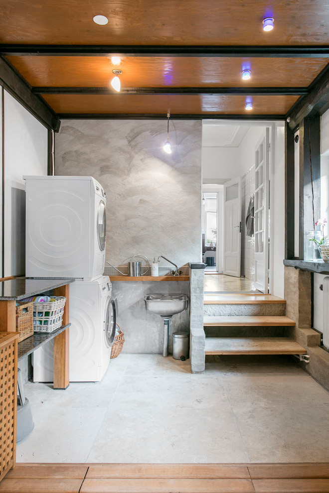 This is an example of a large scandinavian l-shaped laundry room in Malmo with open cabinets, white walls, concrete floors, a stacked washer and dryer and an utility sink.