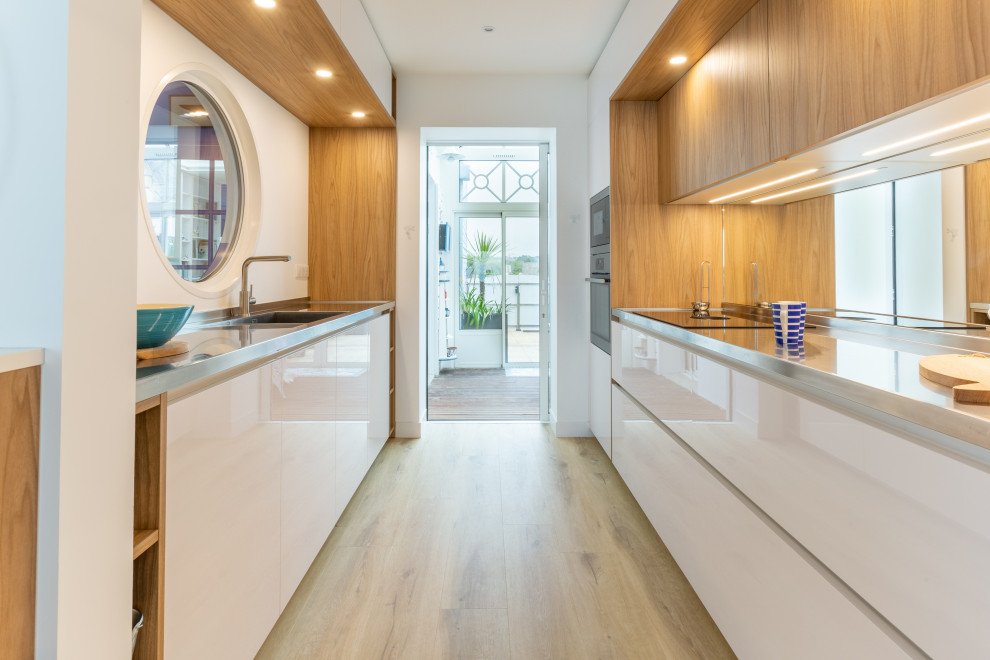 Small beach style galley open plan kitchen in Rennes with an integrated sink, beaded inset cabinets, white cabinets, stainless steel benchtops, mirror splashback, stainless steel appliances, light hardwood floors and timber.