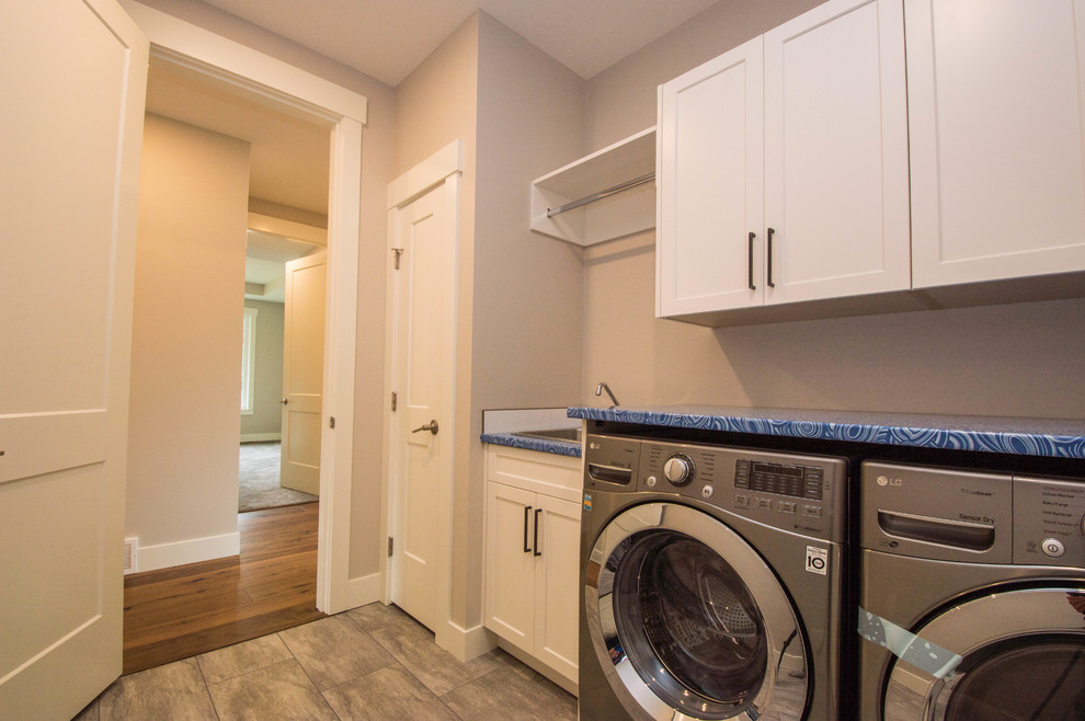 Design ideas for a mid-sized transitional single-wall dedicated laundry room in Vancouver with a drop-in sink, recessed-panel cabinets, white cabinets, laminate benchtops, grey walls, porcelain floors, a side-by-side washer and dryer, brown floor and blue benchtop.