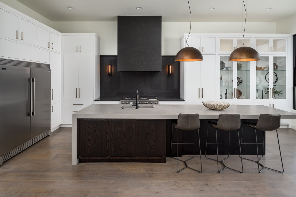 This is an example of a contemporary l-shaped kitchen in Phoenix with flat-panel cabinets, quartz benchtops, stone slab splashback, stainless steel appliances, medium hardwood floors, an undermount sink, white cabinets, black splashback, with island, brown floor and grey benchtop.