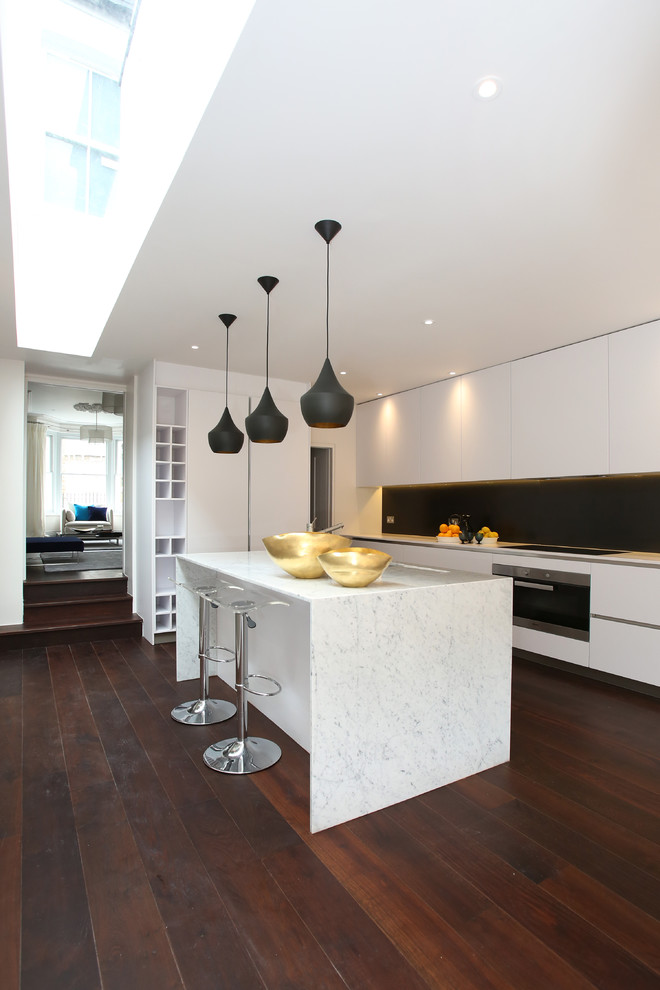 Contemporary kitchen in London with flat-panel cabinets, white cabinets, black splashback, stainless steel appliances, dark hardwood floors, with island and marble benchtops.