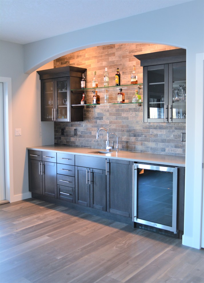Mid-sized transitional single-wall wet bar in Calgary with an undermount sink, glass-front cabinets, dark wood cabinets, solid surface benchtops, multi-coloured splashback, stone tile splashback, medium hardwood floors and grey floor.